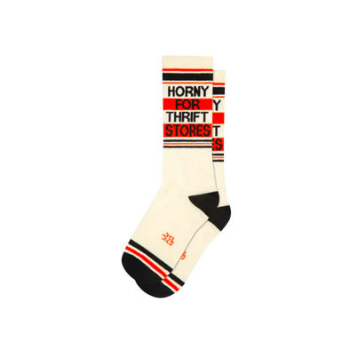 White socks with black and orange stripes and text "Horny For Thrift Stores". Seek out the vintage finds of your dreams with this statement piece. Crafted from a blend of cotton, nylon, and spandex, these unisex socks are designed to fit most and are proudly made in the USA. Slide them on and get ready to hunt for some hidden gems!